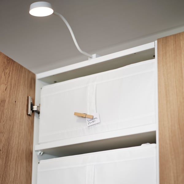 The top shelf of a PAX/TONSTAD white/oak veneer wardrobe holds a white SKUBB storage case. An YTBERG cabinet light is above.