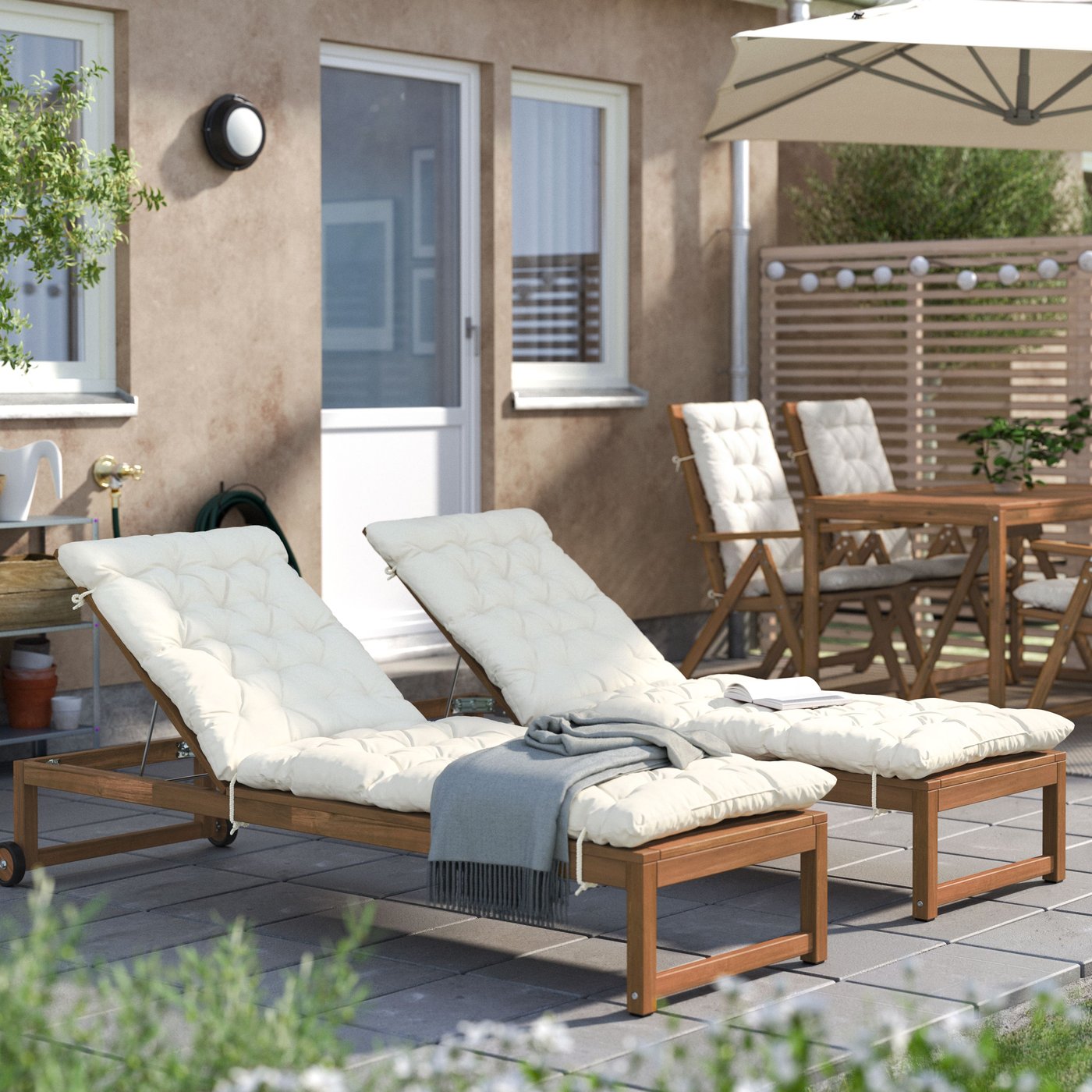 Dvije drvene ležaljke s bijelim jastucima stoje na terasi ispred kuće, uz mali pomoćni stol s dekicom; u pozadini su blagovaonski stol sa stolicama, suncobran i drvena ograda.