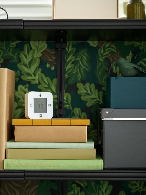 Close-up of bedroom corner with floral-inspired wallpaper, wall shelves, books, boxes with lids, clock with thermometer.