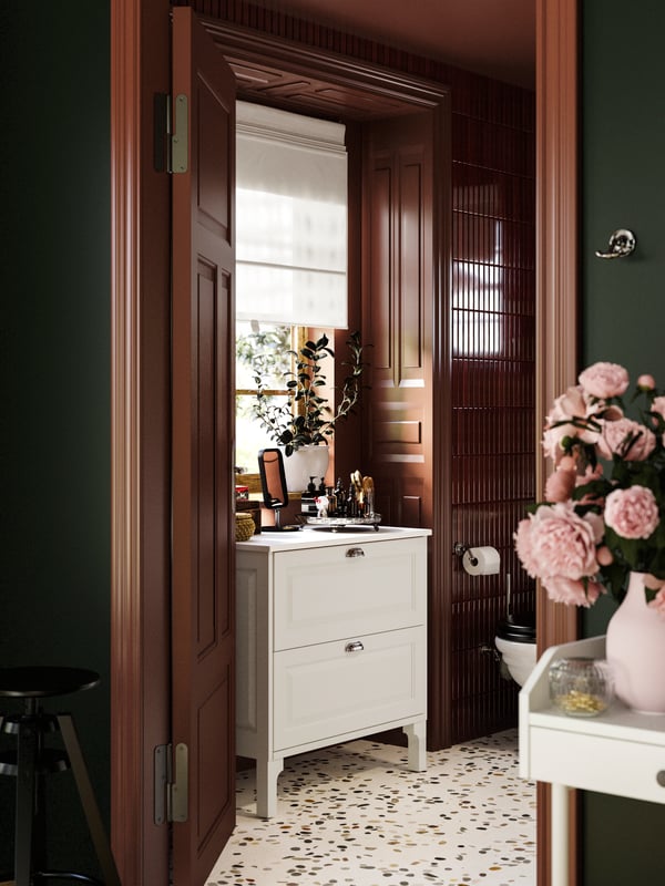 Une salle de bains avec un meuble lavabo à tiroirs TÄNNFORSEN, un vase de fleurs et un mélange de carreaux bourgogne et d’un sol en terrazzo.