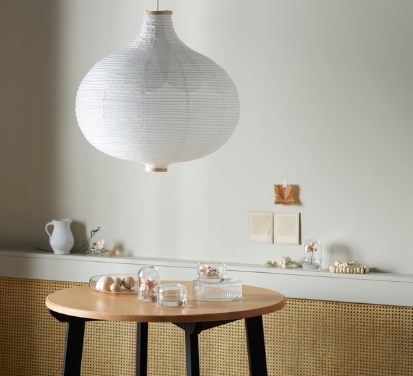 A large RISBYN paper pendant light hangs above a circular wooden coffee table with clear glass vases and dishes. 