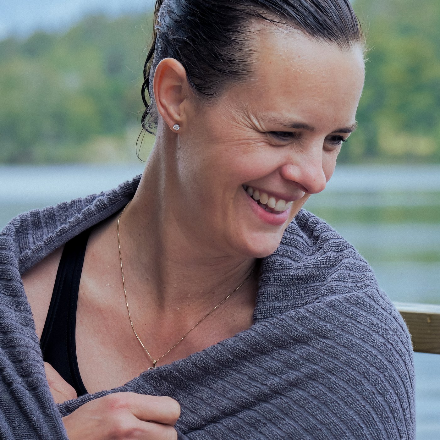 A smiling woman, wrapped in a VÅGSJÖN towel after a swim in a lake.