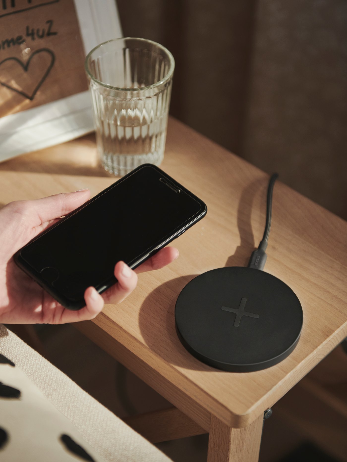 A hand holds a smart phone above a black circular LIVBOJ wireless charger pad that is on a wooden bedside table.