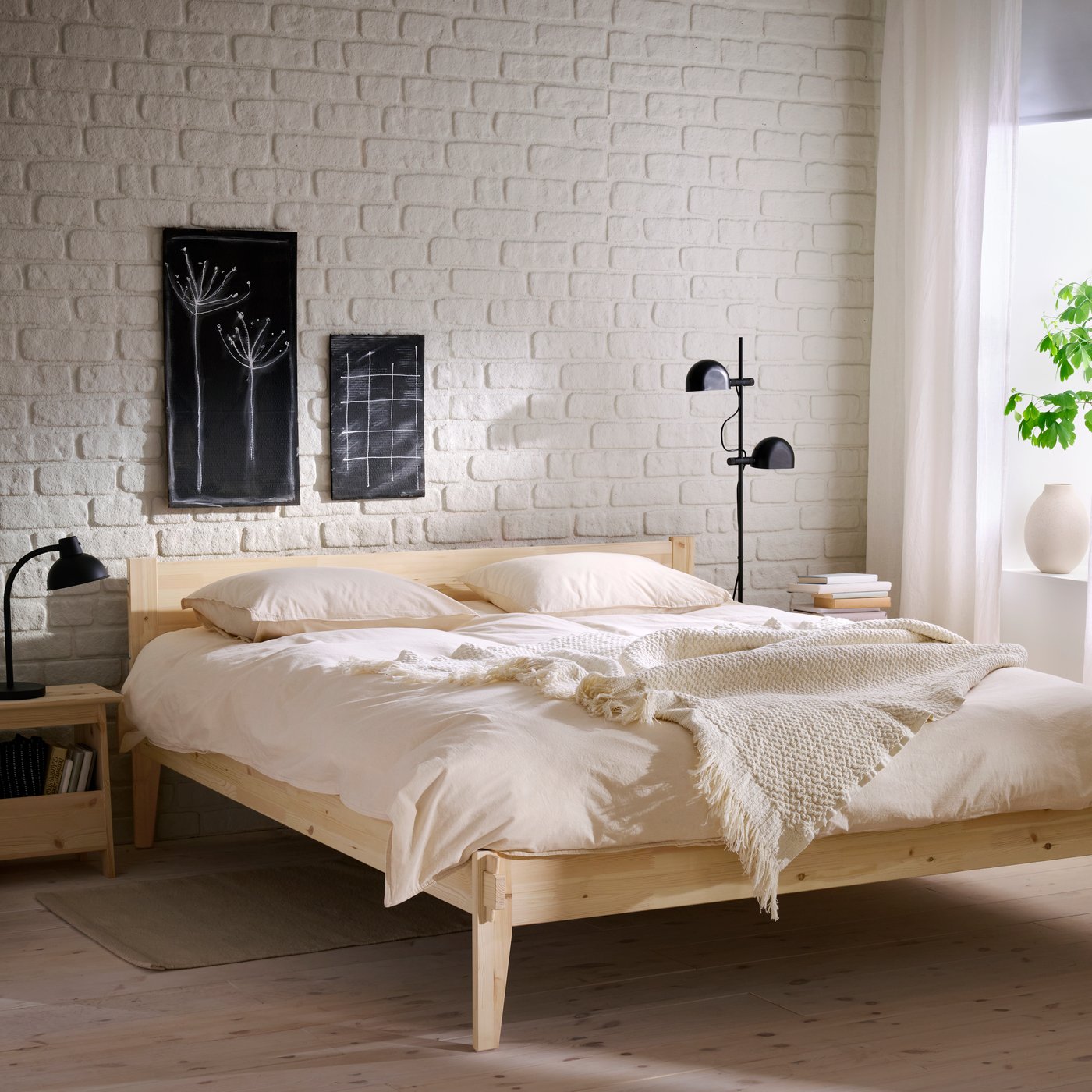 A bedroom with a white brick wall, a wooden TAXÅSKLINT bed with a soft beige duvet and pillows and a throw and black lamps.