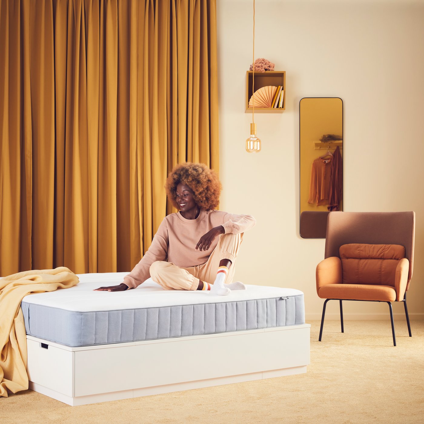 Una ragazza è seduta su un letto con materasso VESTERÖY. Alle sue spalle una tenda arancione e uno specchio con davanti una poltrona.