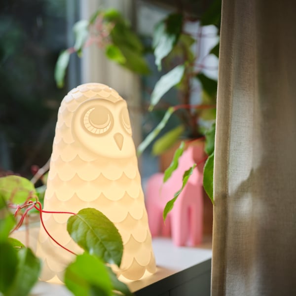 SOLBO LED table lamp shaped as a white owl standing in a window, surrounded by plants and the curtain.