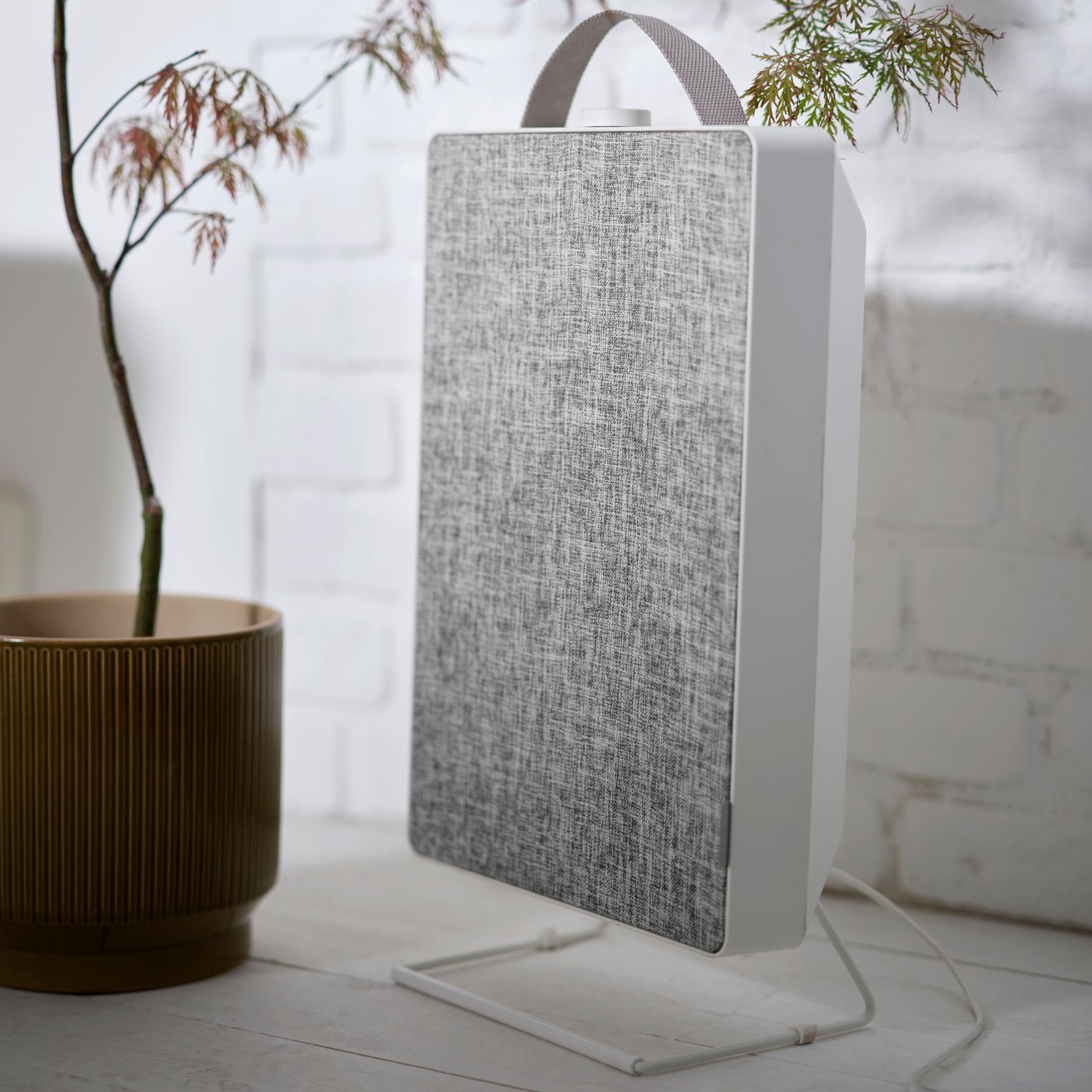 A white FÖRNUFTIG air purifier next to a plant in front of a white painted brick wall.