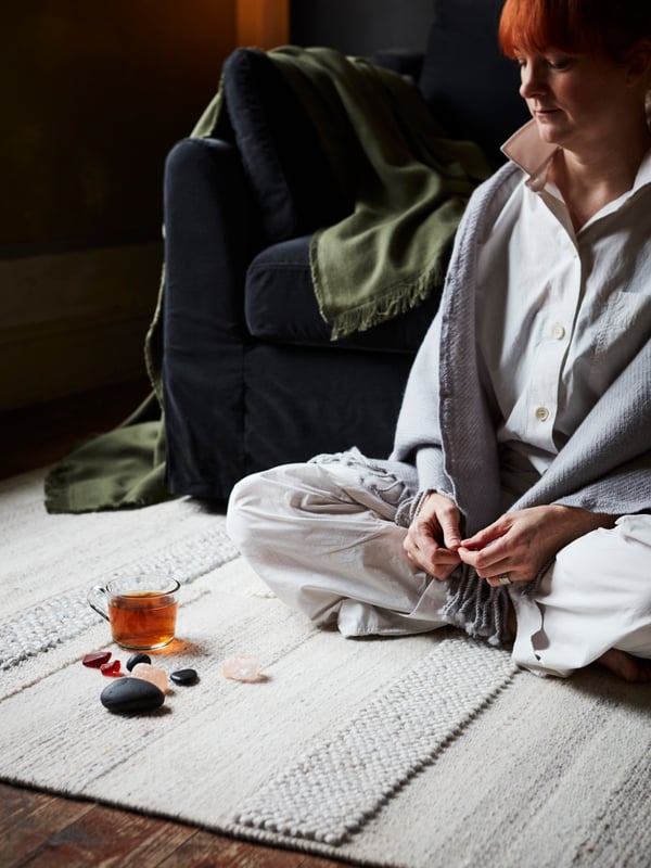 A woman sits cross-legged on a handwoven beige BRÖNDEN rug, beside a FÄRLÖV dark gray sofa with cushions and a throw.