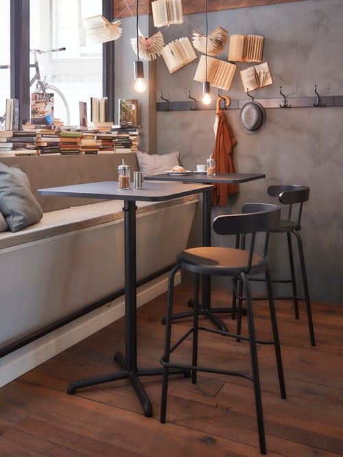 O sală de mese cu 2 mese de bar pătrate din oțel și plăci aglomerate amplasate lângă 2 scaune de bar gri.