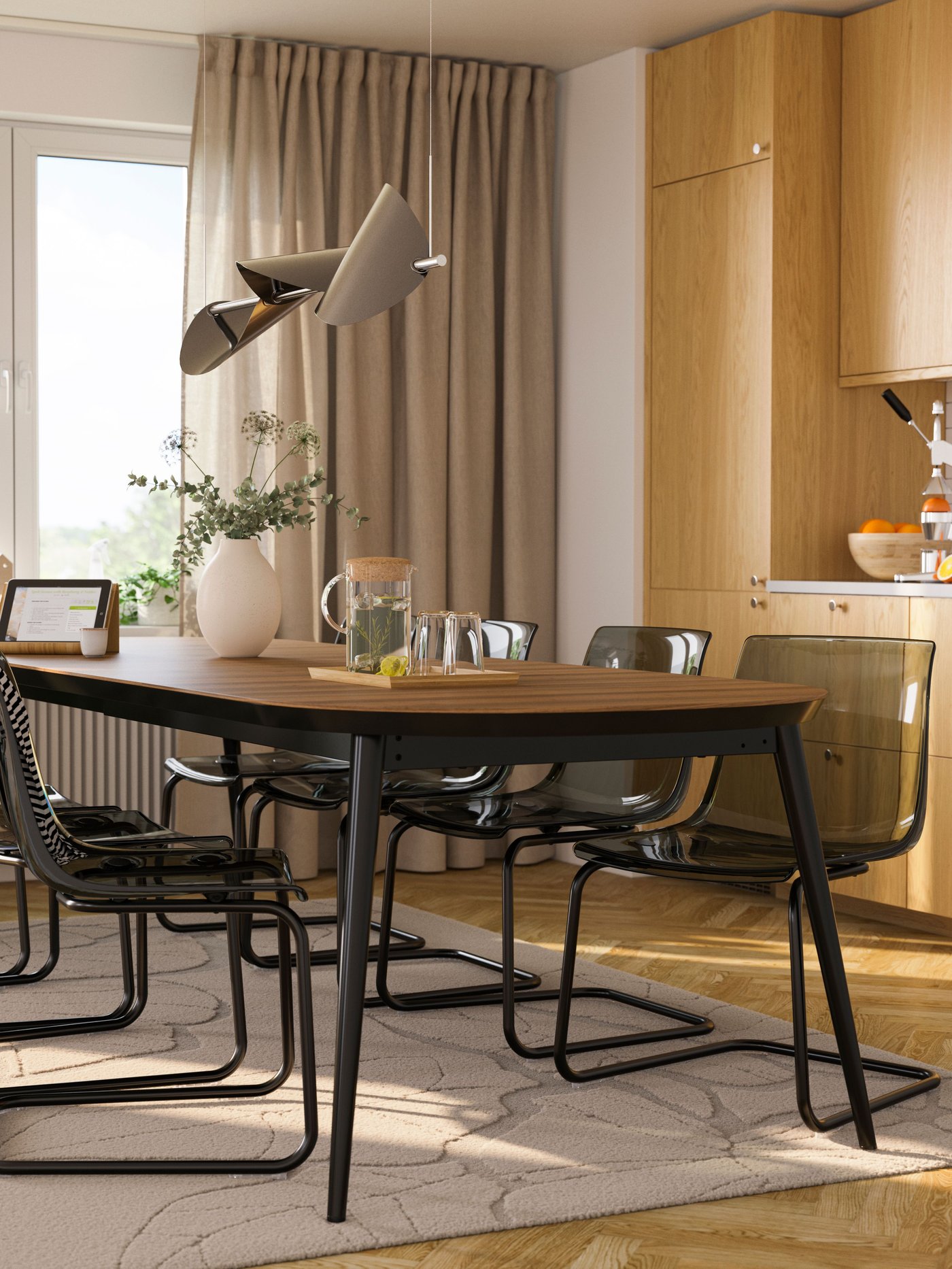 Une salle à manger chaleureuse présente une table ovale en bois foncé entourée de chaises noires à piètement luge, un élégant luminaire suspendu, un vase en céramique fleuri et une cuisine en bois clair en arrière-plan.