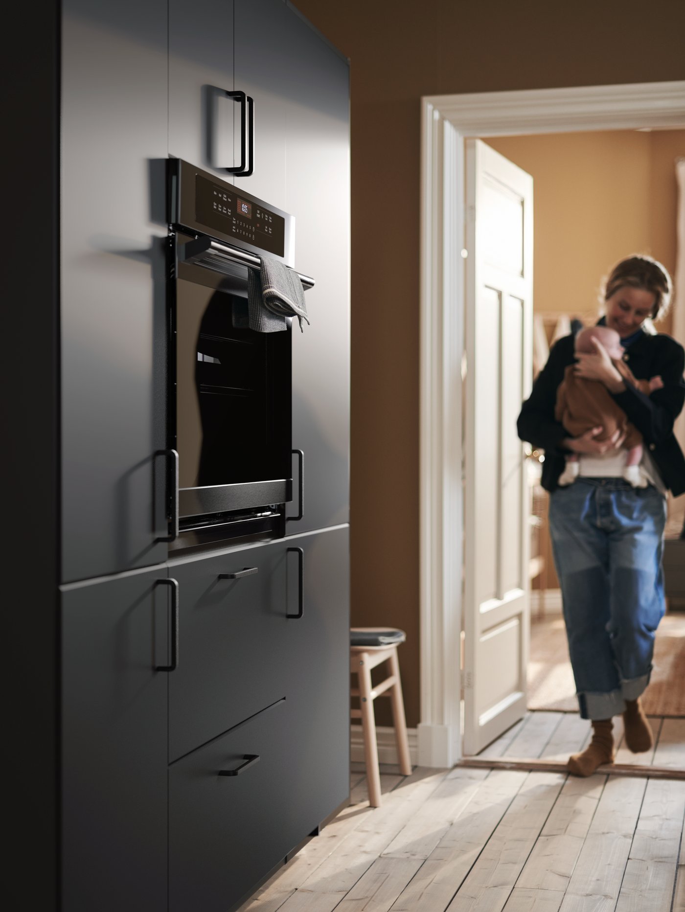 A mother cradles her baby as she enters the kitchen, approaching a wall of cabinets with KUNGSBACKA fronts.