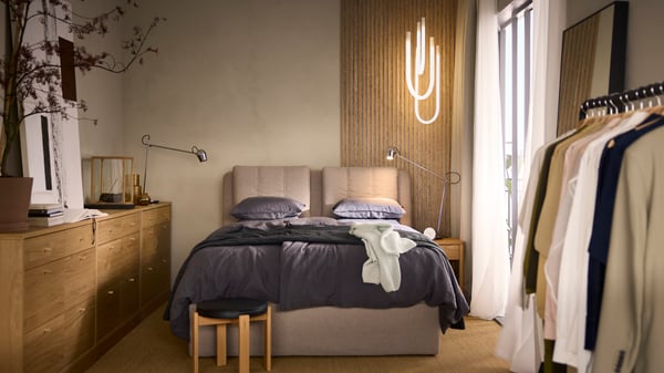 Ein LILLFJÄLLET Boxspringbett mit Bettkasten ist mit Bettzeug in dunkelgrauer Bettwäsche bezogen. Es steht an einer beigefarbenen Wand. Darüber hängt eine VARMBLIXT LED-Hängeleuchte.
