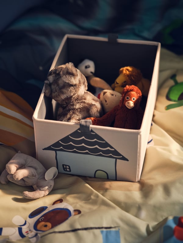 A beige BARNDROM box holding soft toys including a LILLEPLUTT cat and a DJUNGELSKOG orangutan on top of a bed.