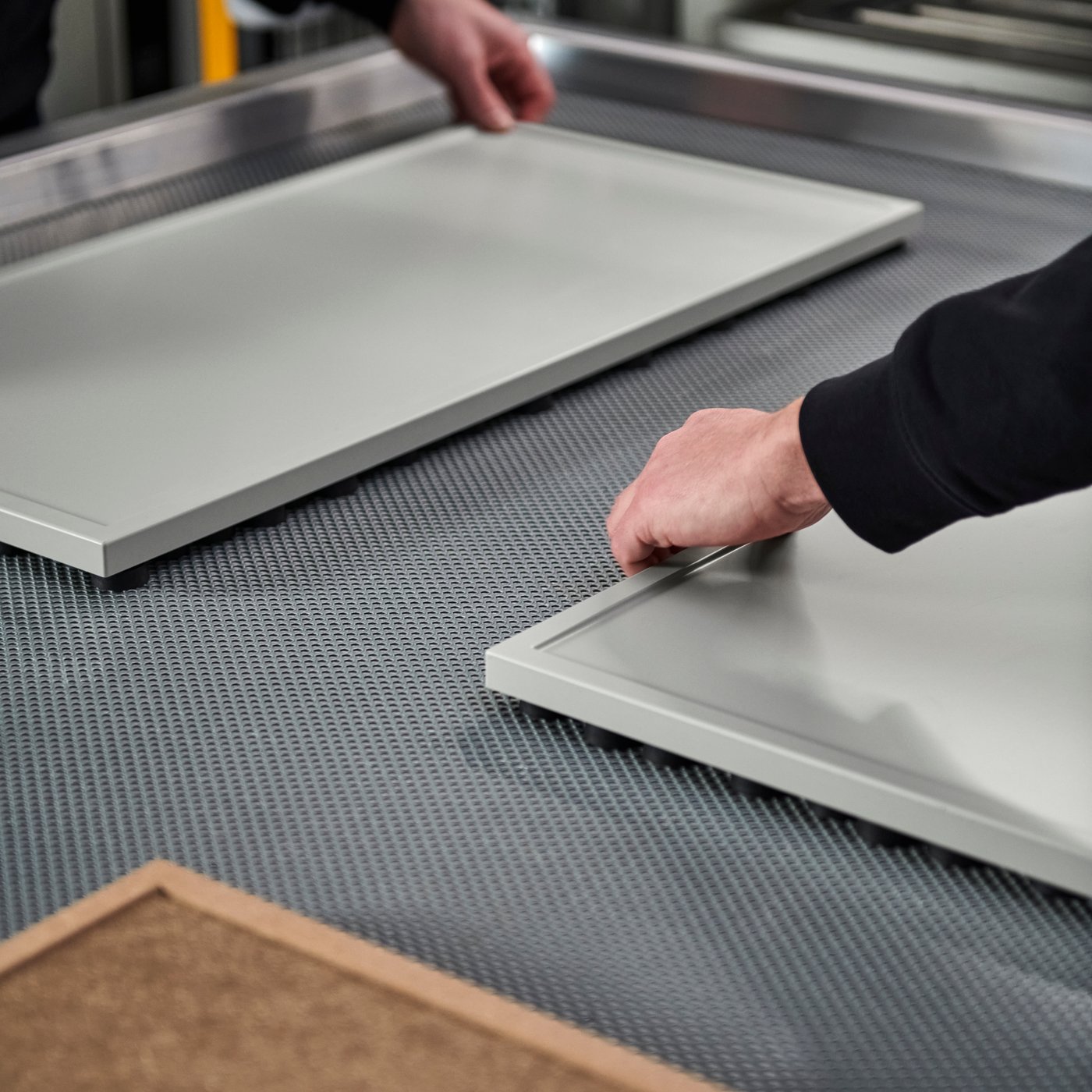 A close-up of unfinished and finished ASPUDDEN light grey door fronts being inspected on a membrane press machine’s tray.