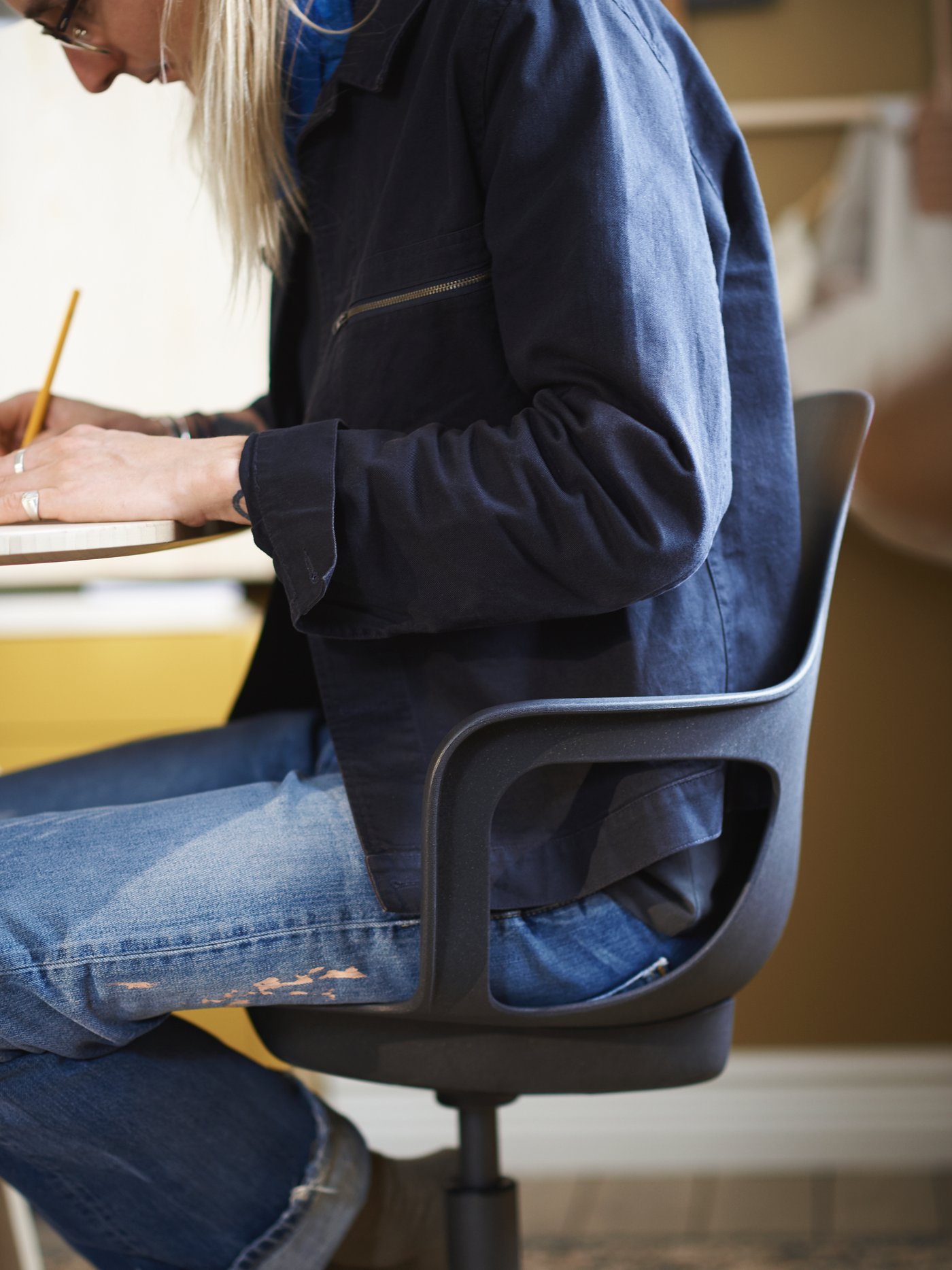 A woman sitting on an ODGER swivel chair in anthracite, by a table, writing on a pad with a pencil.