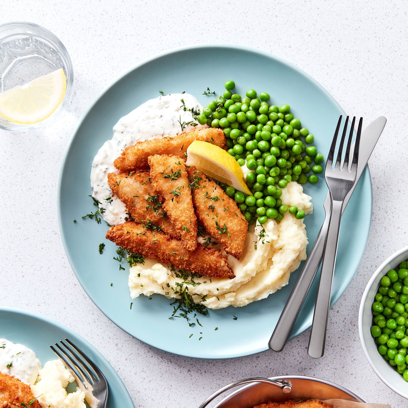 Cinq bouchées de poisson pané dorées servies avec une purée de pommes de terre, des petits pois et un dip dans une assiette bleue.