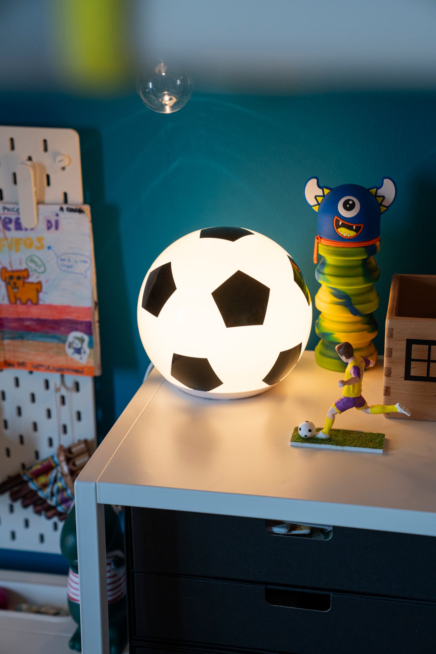 Lampada da tavolo a forma di pallone da calcio accesa su mobile in cameretta, accanto a piccoli giocattoli colorati.