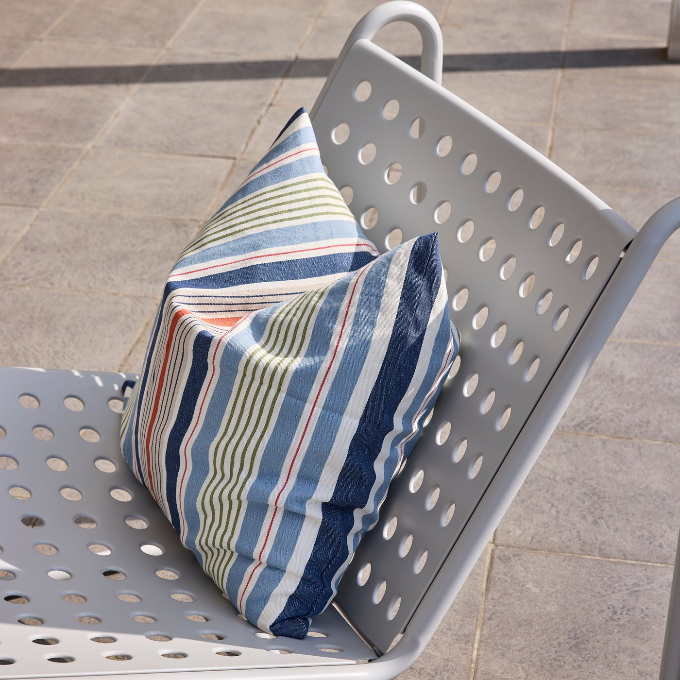 Gestreept kussen in blauw en wit op een geperforeerde witte metalen stoel op een terras.
