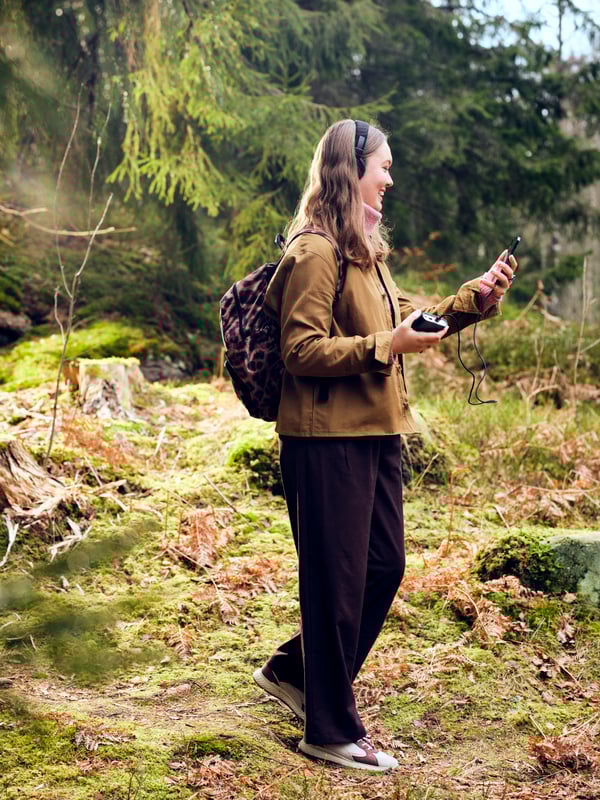 A smiling, leisurely dressed, young woman walks in forest terrain, holding a phone attached to a VARMFRONT power bank.