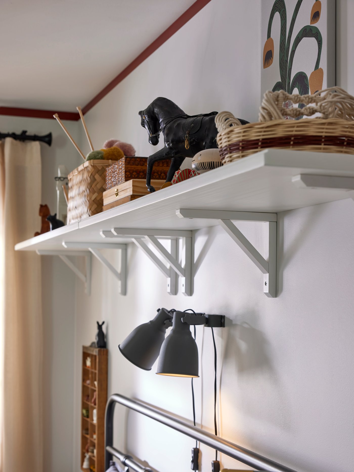 En vit hylla med korgar och dekorationer är monterad på väggen ovanför en säng. Under hyllan sitter två HEKTAR väggspotlights.
