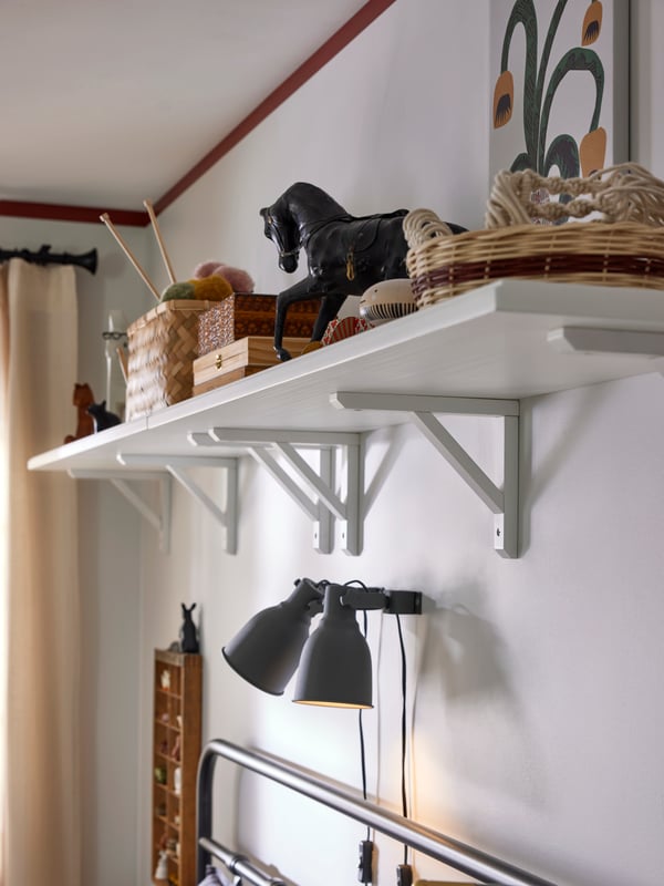 White shelf with baskets and decoration on, mounted on the wall over bed. Two HEKTAR wall spotlights underneath.