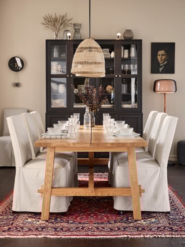 IKEA BERGMUND upholstered dining chairs with long covers in an off-white colour, in front of a large oak dining table, an oriental rug, a dark brown display cabinet and rattan ceiling lights.