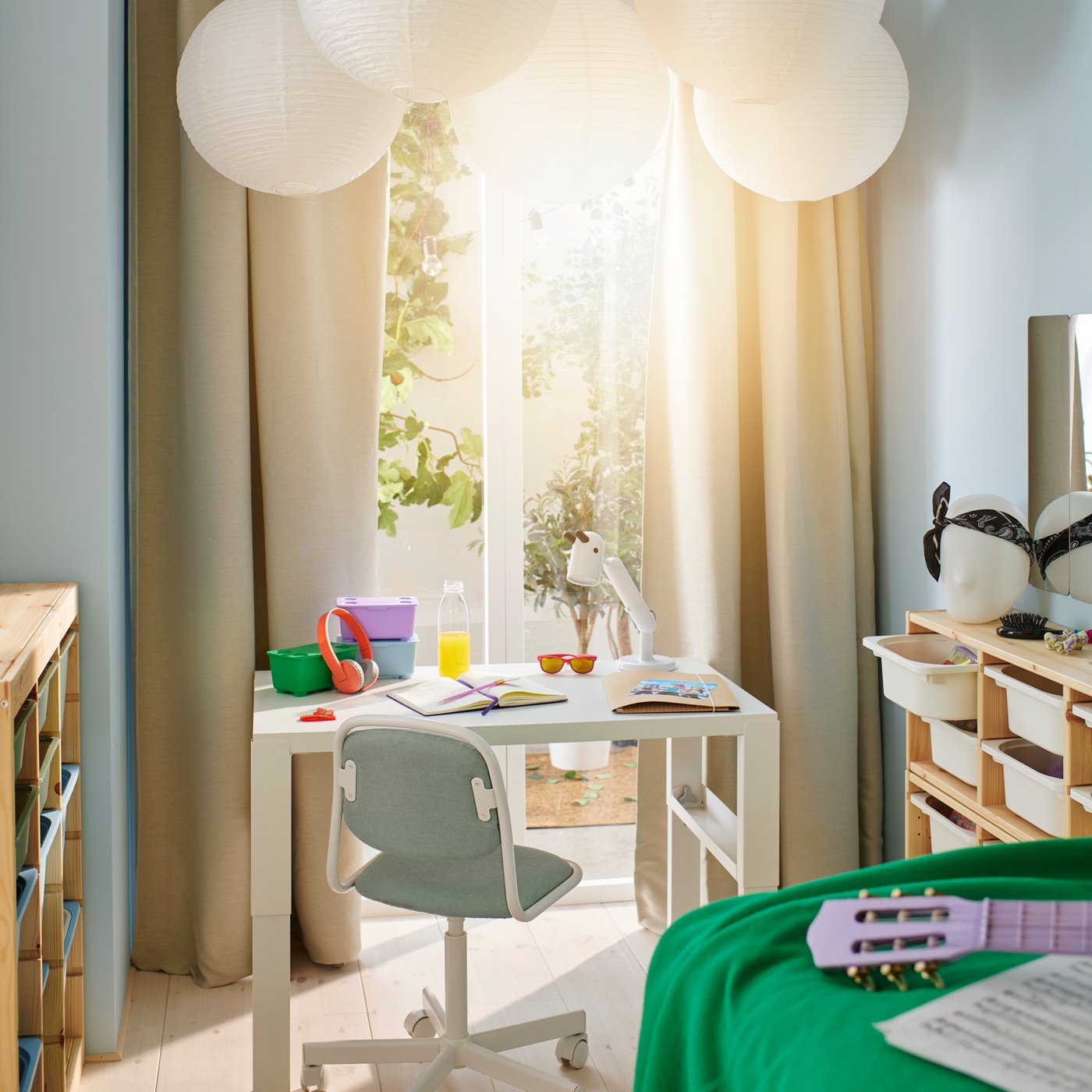 A white PÅHL desk in a bright sunny room that has green, lilac and blue GLIS boxes and a white KRUX LED work lamp on top.