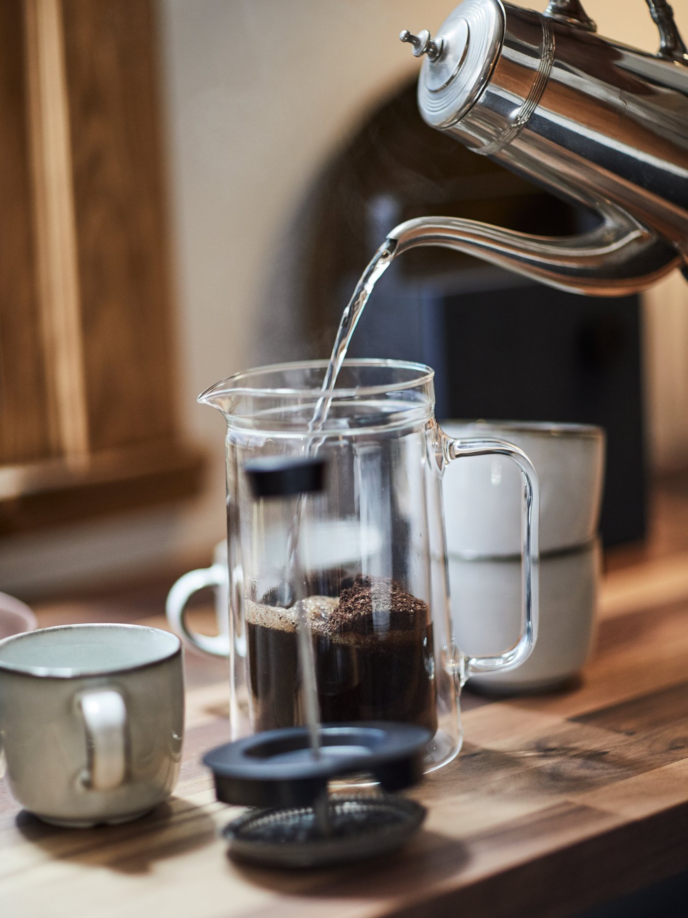 A silver jug pouring water over ground coffee beans in an EGENTLIG coffee/tea maker with GLADELIG mugs to the left and right.