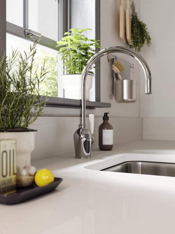 A white kitchen worktop, a chrome-plated kitchen mixer tap, a sink in stainless steel and some herbs by a window.