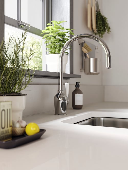 A white, custom made worktop, two potted plants, a kitchen mixer tap, and a grey container on the wall.