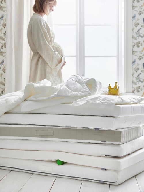 A pregnant woman in a white room between a window and a pile of cot mattresses including DRÖMMANDE, PLUTTIG and KRUMMELUR.