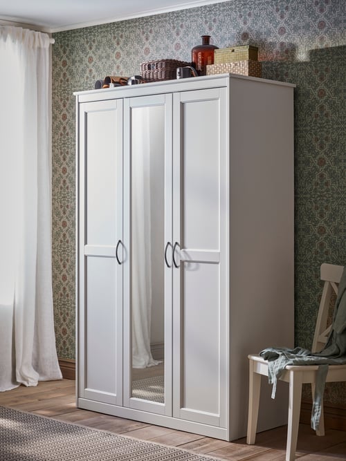 A SONGESAND white wardrobe has a mirrored door with boxes, paper rolls, baskets and a vase on top by a chair and a window.