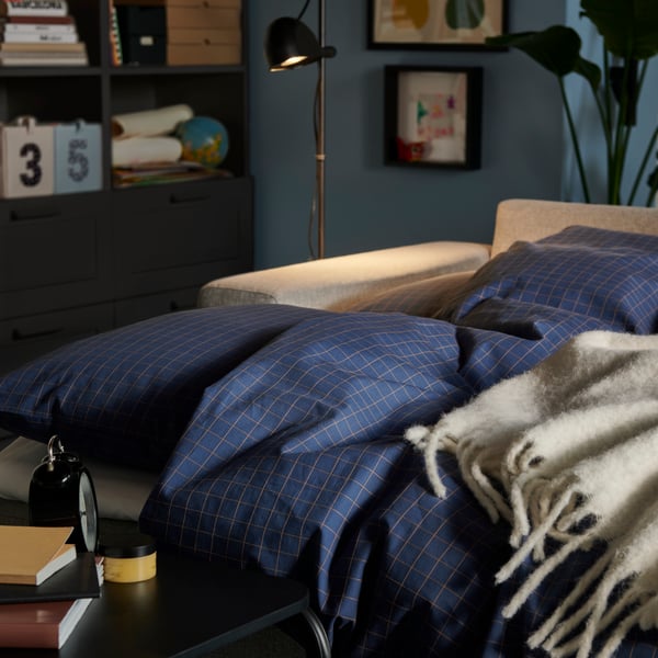 A softly lit living room with a BÅRSLÖV sofa-bed made with DOFTAKLEJA bed linen and an OLSERÖD side table by the bed.