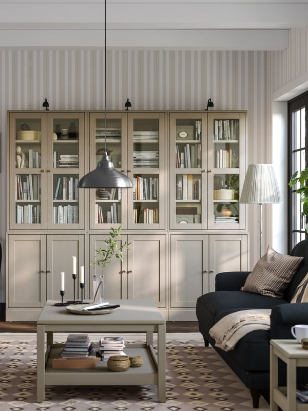 Ein HAVSTA Kombination in graubeige aus 3 Vitrinenschränken, bei denen die Vitrinenaufsätze leicht eingerückt sind steht an einer gestreiften Wand. Jeder Vitrinenschrank ist beleuchtet, damit man Bücher und Deko gut sehen kann. Davor steht ein Sofa und der passende HAVSTA Couchtisch.