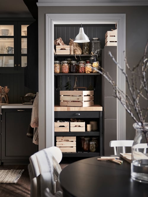 A kitchen pantry with a kitchen island with boxes, shelves with boxes and jars, a pendant lamp, and a fridge.