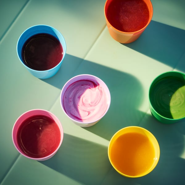 Sur une table vert clair placée au soleil, six chopes pour enfants KALAS en bleu, orange, rose, vert et jaune, remplies de jus colorés.