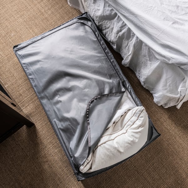 A gray SKUBB storage case with pillows and duvets in it on the floor next to a bed.