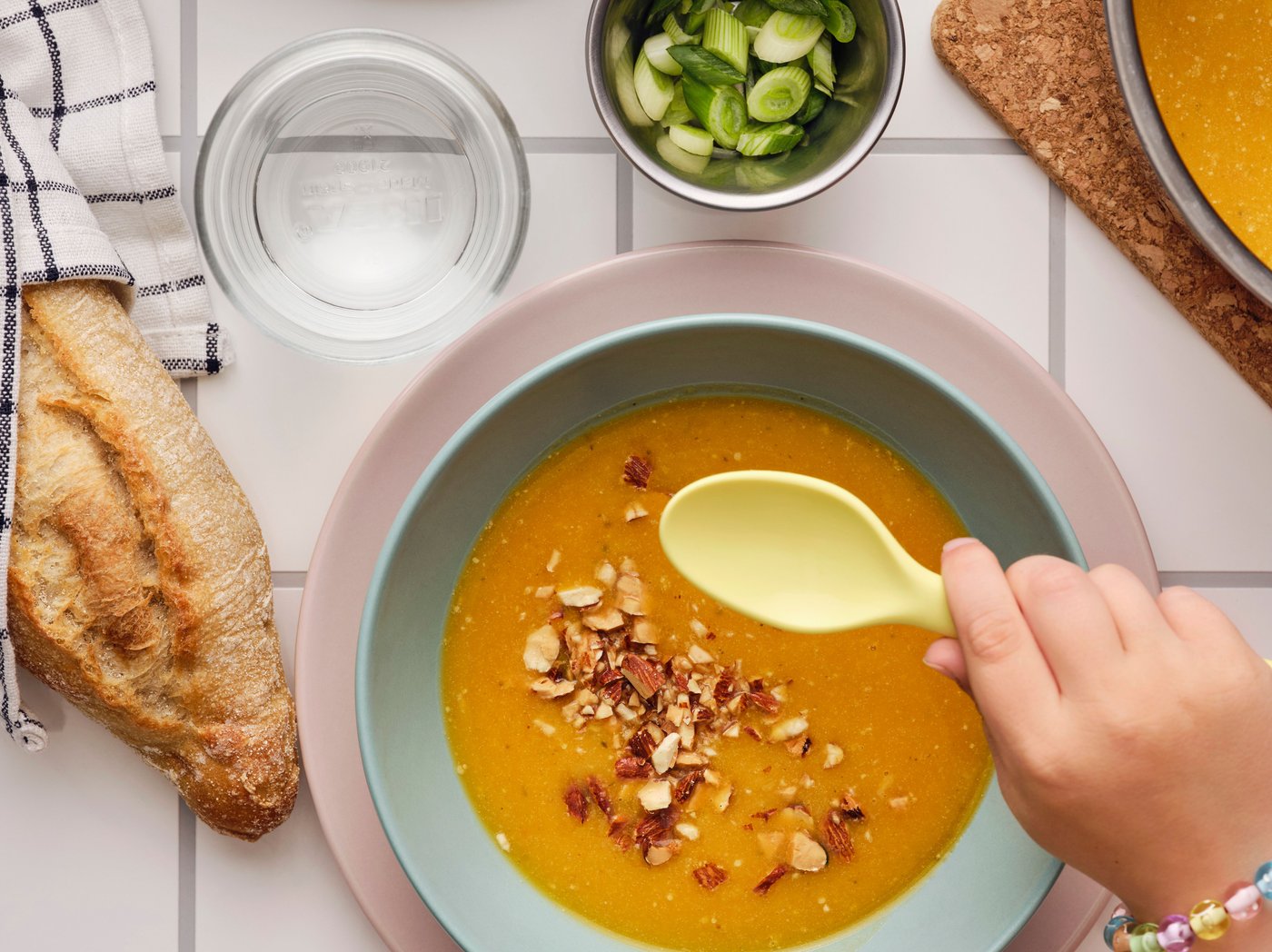 A hand holding a spoon over a bowl of pumpkin soup garnished with MUNSBIT roasted almonds, with bread and herbs on the side.