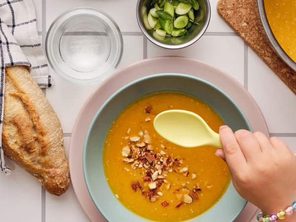 A hand holding a spoon over a bowl of pumpkin soup garnished with MUNSBIT roasted almonds, with bread and herbs on the side.