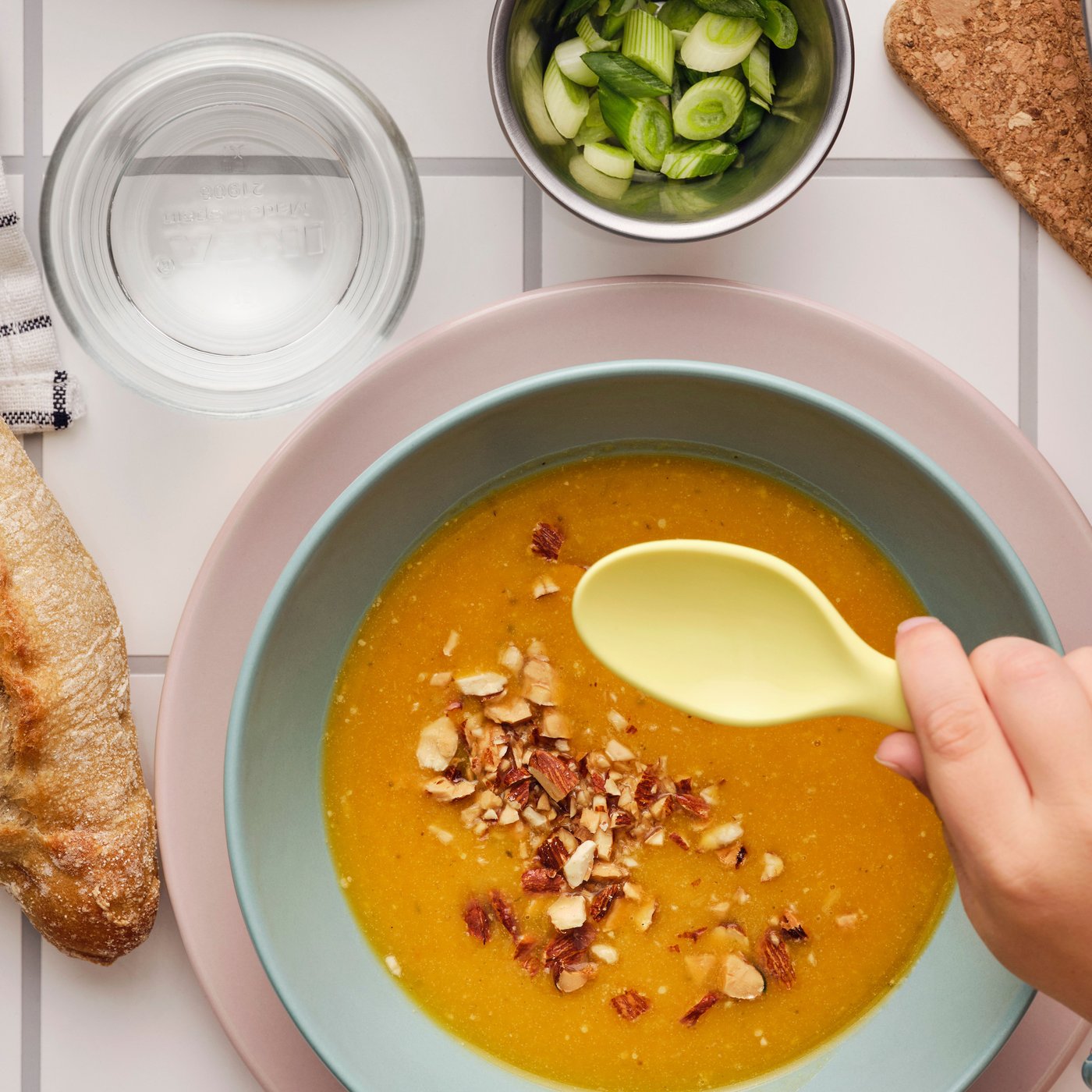 Main tenant une cuillère au-dessus d’un bol de soupe au potiron garnie d’amandes grillées MUNSBIT entouré de pain et d’aromates.
