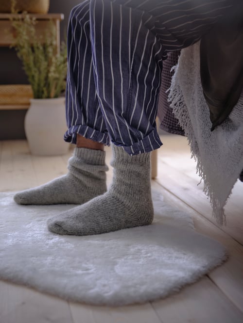 Bedroom with person sitting on a bed frame, with a throw on it, wearing socks and pyjamas, while placing feet on a white rug.