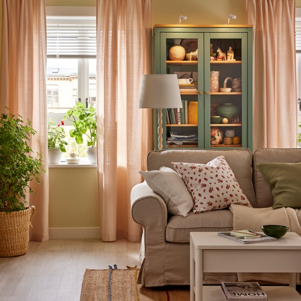 LENDA gardiner i lys pink hænger i to vinduer på hver side af et HEMNES vitrineskab bag en sofa.