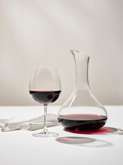 A photo of a dining room that features a red wine glass next to a carafe, both half filled with red wine.