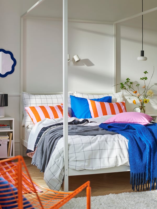 Bedroom with VITARNA four-poster bed frame with colourful bedding, part of bright orange SKÅLBODA armchair in foreground.