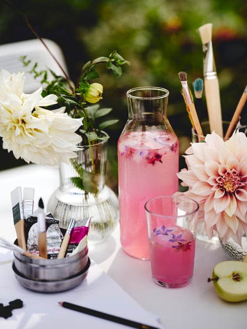 O carafă IKEA 365+ în aer liber cu un dop umplut cu suc, o vază cu flori, un pahar și un set de perii MÅLA.