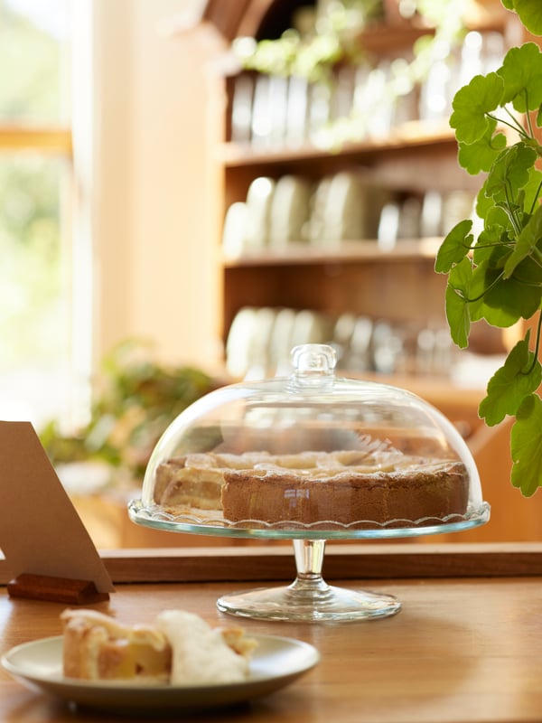 Un'alzata per torte ARV BRÖLLOP con dentro un dolce si trova su una scrivania accanto a un laptop e a un piatto con una fetta di torta.