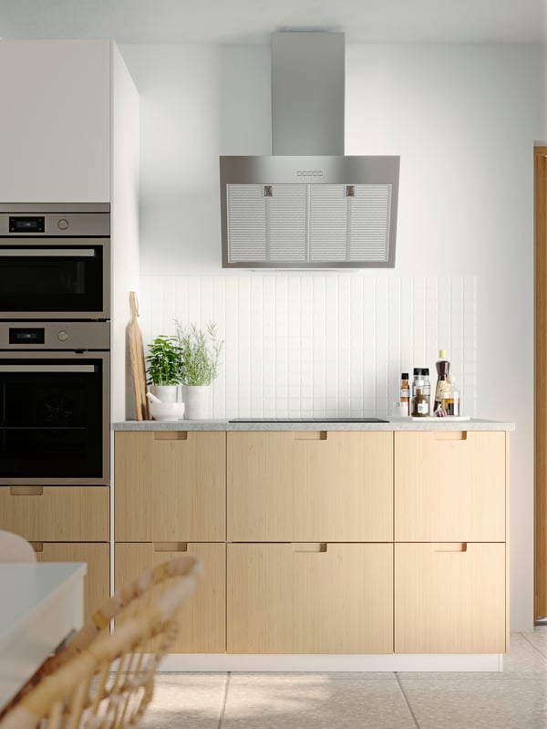 Cuisine avec murs blancs, petit carrelage blanc, façades de cuisine FRÖJERED en bambou et électroménager en acier inoxydable.