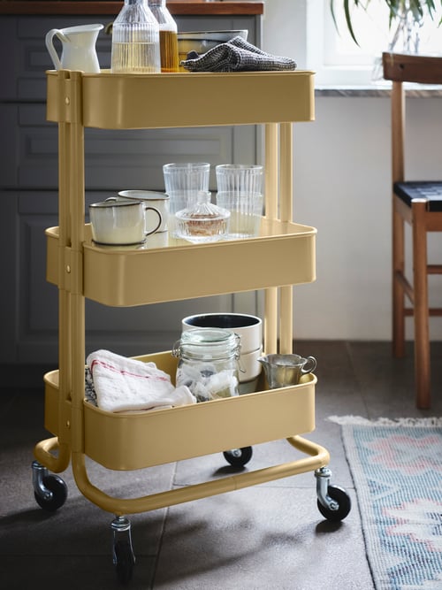 Un coin cuisine dans lequel se trouvent un bocal avec couvercle, une tasse, un vase, une carafe et un bol avec couvercle, le tout placé dans un chariot jaune.