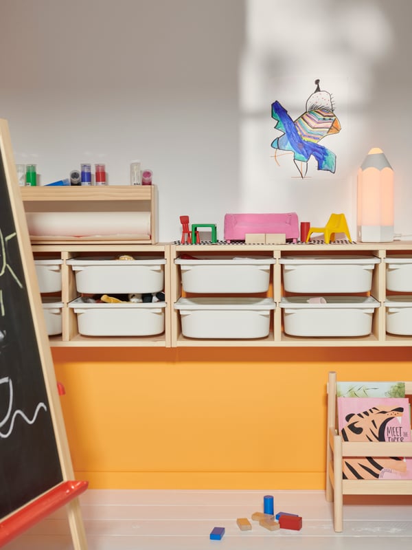 Zwei weiß gebeizte TROFAST Wandaufbewahrungen in Kiefer hängen an einer orange-weißen Wand. In den Aufbewahrungen befinden sich weiße TROFAST Boxen.