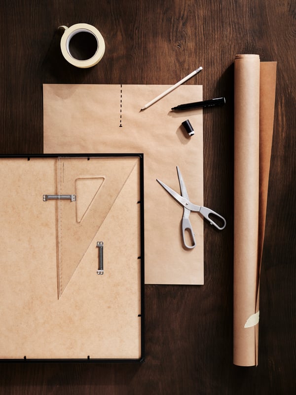 A dark-wood surface with SVÄRDFISK scissors, a paper roll and a measuring tool on and next to a flipside-up RÖDALM frame.